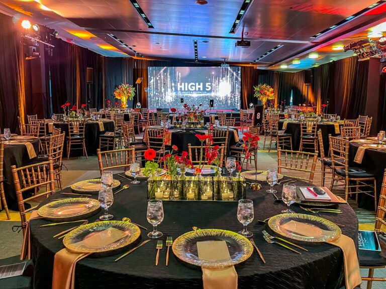 Elegant gala dinner setup by Showorks in a hotel ballroom with vibrant red and orange floral centerpieces, chic bamboo chairs, and an illuminated 'HIGH 5' sign on a grand stage, showcasing their expertise in event organization and design.