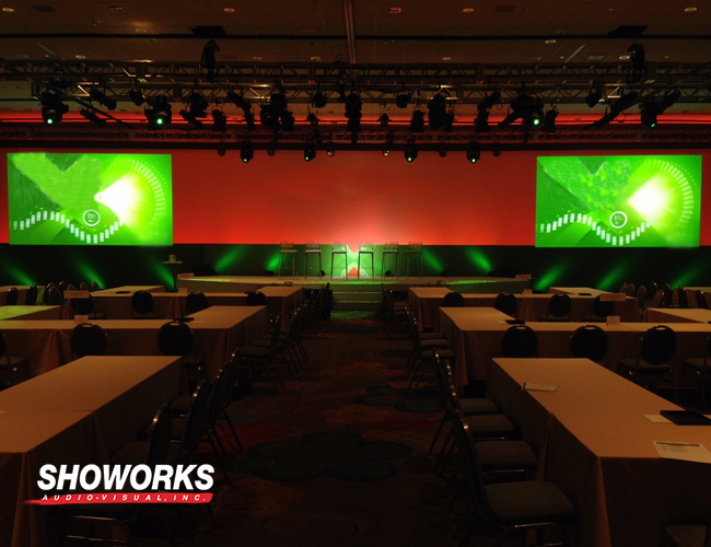 A professionally staged conference event setup by Showorks Audio-Visual, Inc., featuring dynamic green lighting, dual projection screens, and a well-lit stage for speakers.