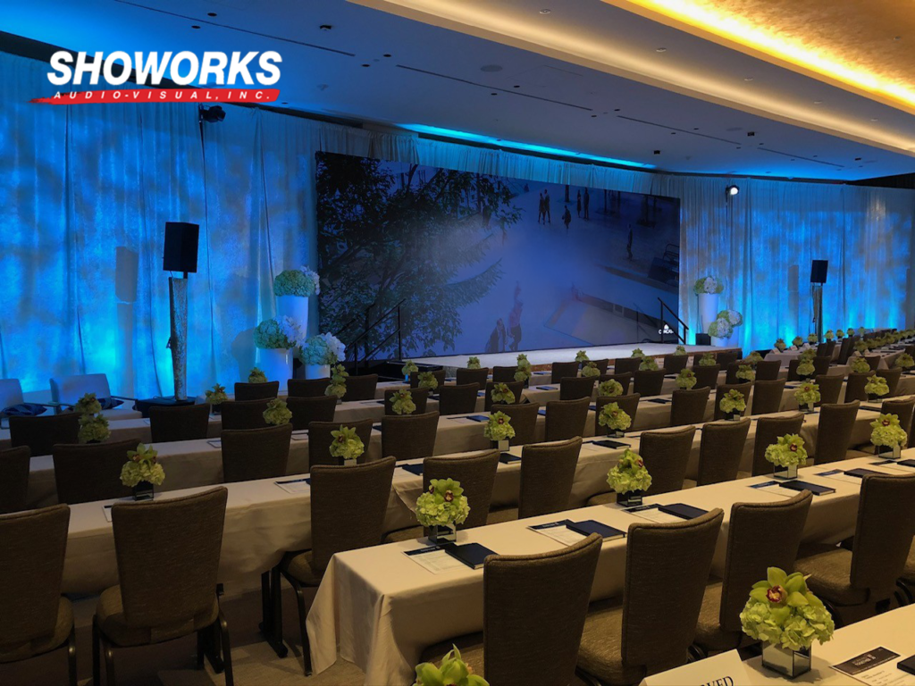 Corporate conference room setup with floral centerpieces, blue ambient lighting, and a large stage backdrop, produced by Showorks AV.
