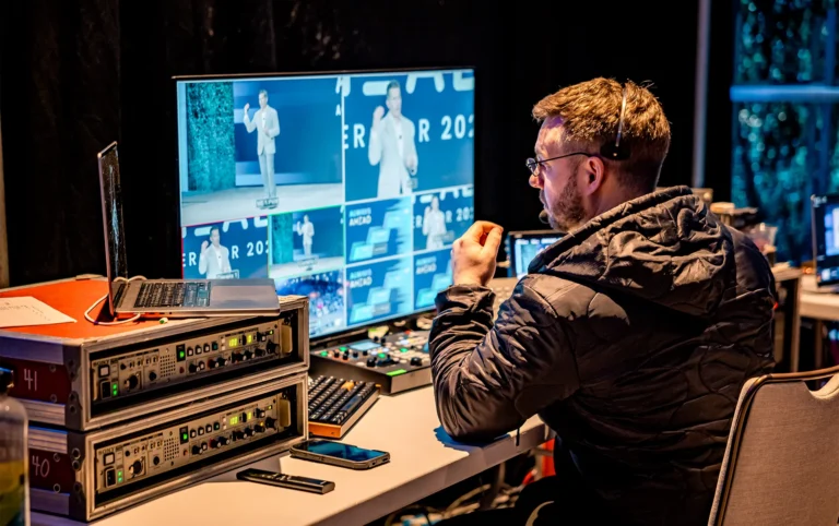 Production manager overseeing live corporate event production services from an AV control station during a conference presentation.