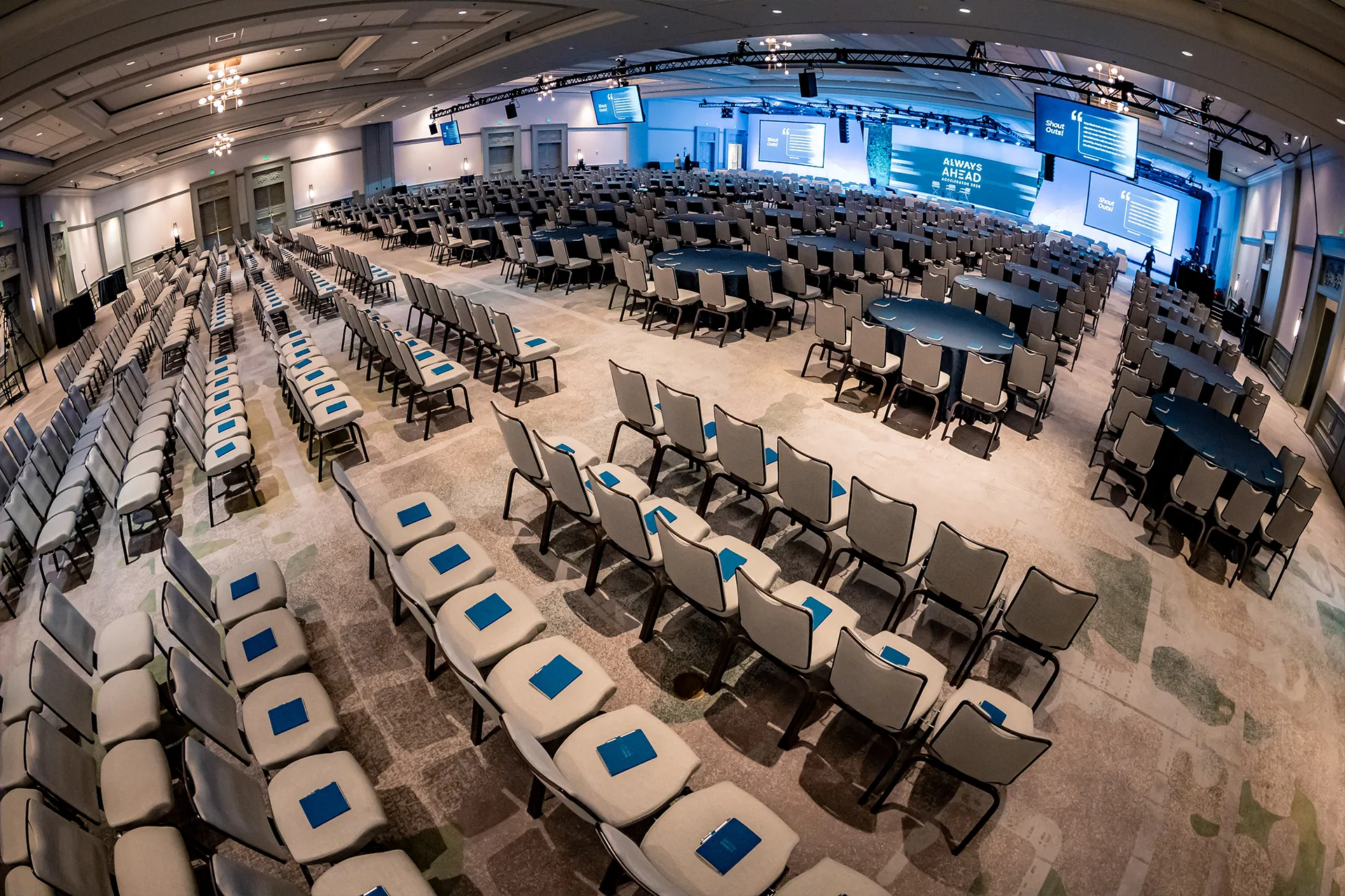An empty conference room is set with rows of chairs, tables, stage lighting, and presentation screens before a corporate event.