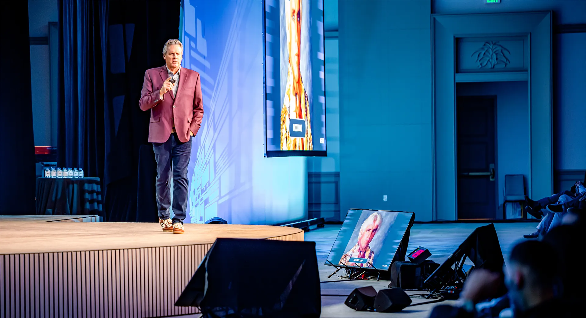 A presenter rehearses on a lit stage with confidence monitors and AV equipment before a corporate event.