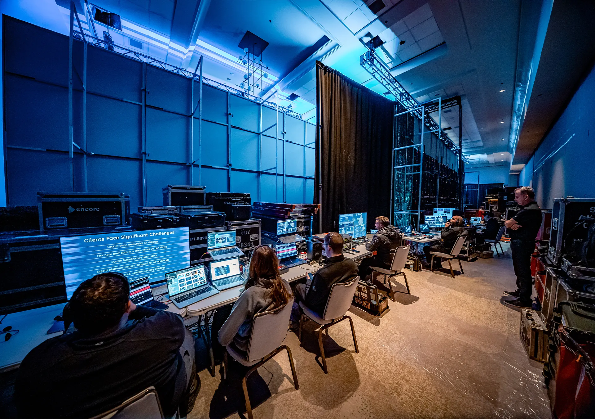 Production team monitoring multiple screens and laptops backstage, with a slide on client challenges displayed, showing how AI in AV production helps address complex technical and operational demands.
