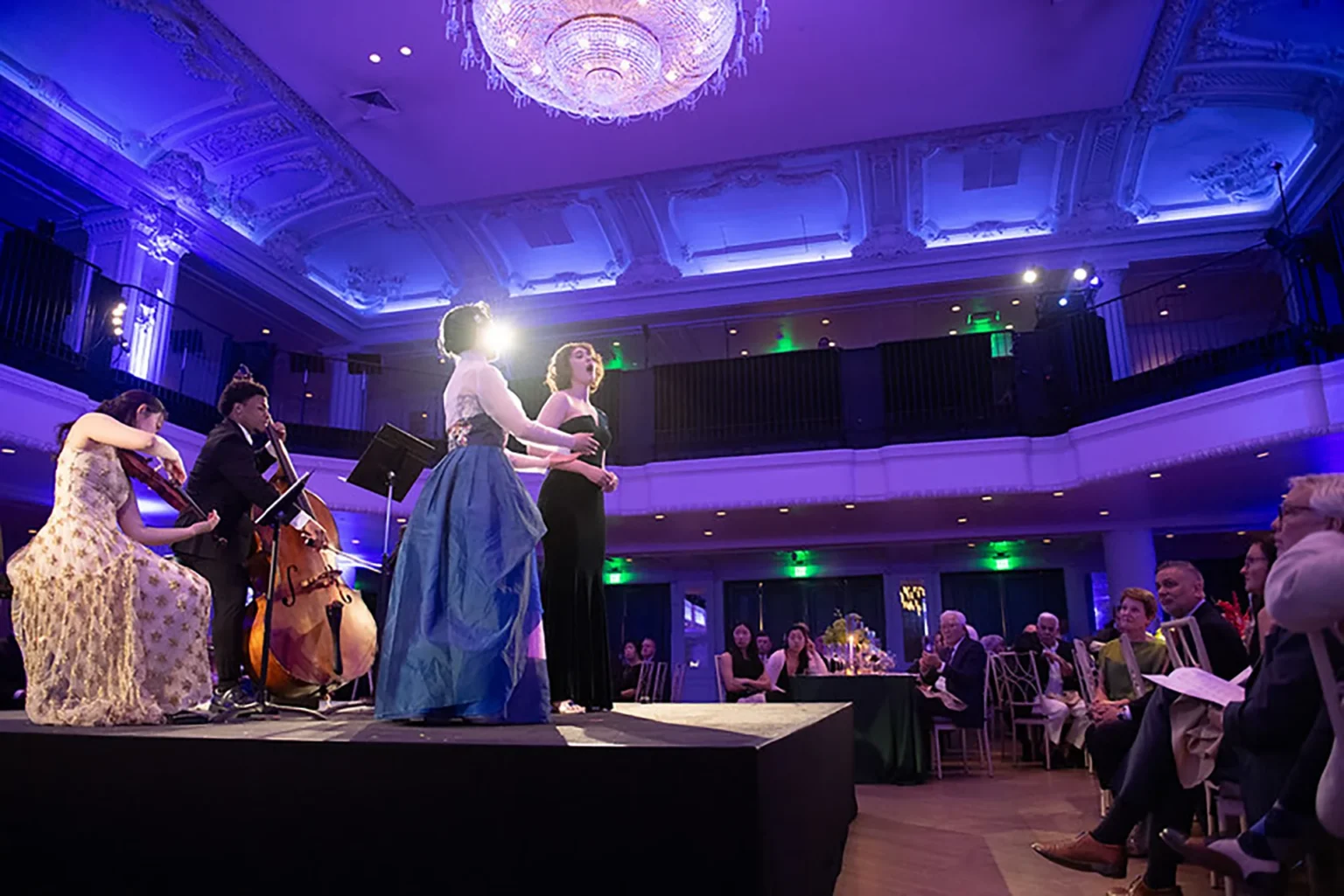 A vocalist and string musicians perform onstage at a gala beneath blue and purple event lighting.