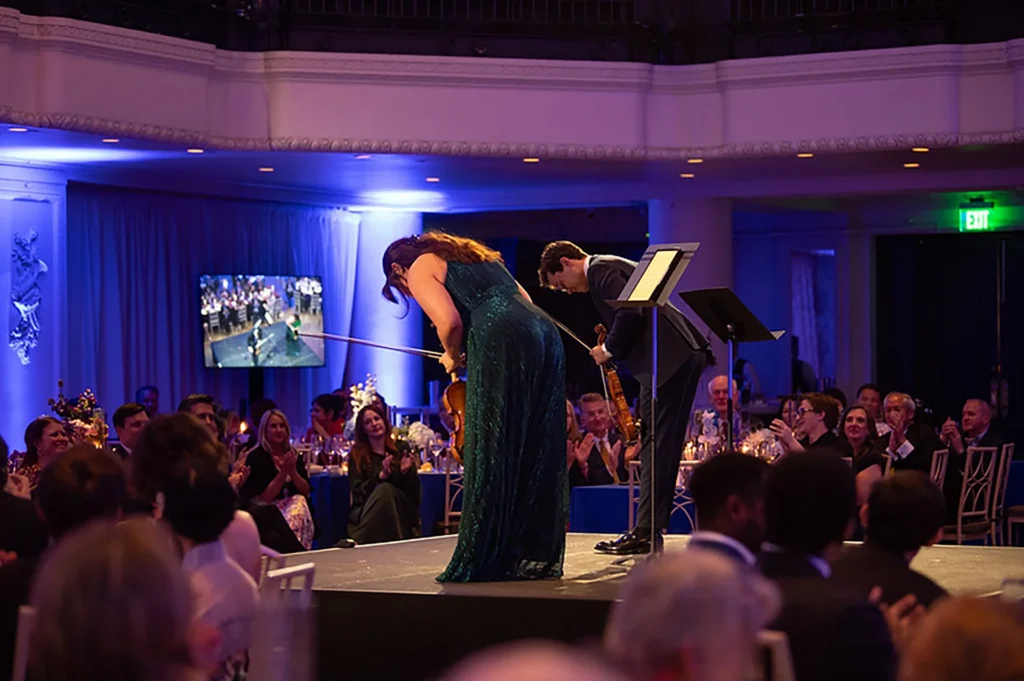 A singer and violinist perform onstage as gala guests applaud under blue lighting.