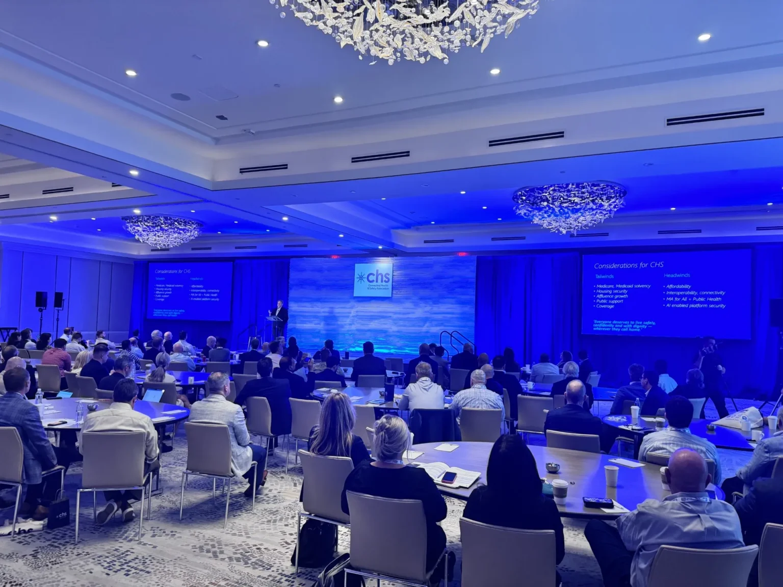 Audience seated at round tables listening to a speaker presenting on a stage with blue lighting.