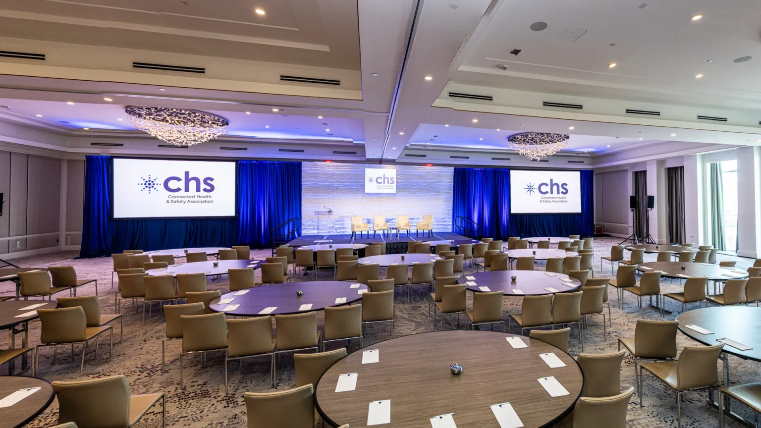 Conference room with round tables and chairs facing a stage with four seats and Connected Health & Safety Association screens.